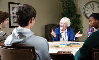 A senior citizen talks to three high schoolers at a table.