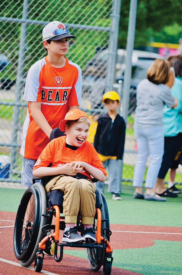 A boy pushes another boy in a wheelchair at an East Metro Miracle League game