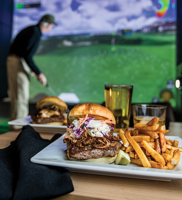 In the foreground, a burger and fries from Birdi Golf. In the background, a man plays indoor golf.